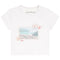 A white short-sleeve crewneck t-shirt by Katie J, featuring a vintage postage stamp graphic on the front with a coastal landscape, shells, and a stamp cancellation mark.