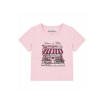 A light pink short-sleeve t-shirt for kids by Katie J, featuring a graphic print of a Parisian-style boutique with red and white striped awning, pink flowers, and a heart on the front.