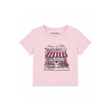 A light pink short-sleeve t-shirt for kids by Katie J, featuring a graphic print of a Parisian-style boutique with red and white striped awning, pink flowers, and a heart on the front.