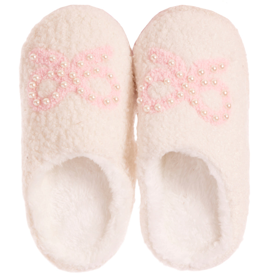 A pair of fluffy off-white slip-on slippers with a light pink bow design embellished with small white pearls on the top, shown against a plain white background.