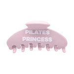 A light pink hair claw clip with the words 'PILATES PRINCESS' printed in bold white capital letters across the front, set against a plain white background.