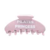 A light pink hair claw clip with the words 'PILATES PRINCESS' printed in bold white capital letters across the front, set against a plain white background.