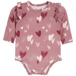 A dusty rose pink long-sleeve baby bodysuit with an all-over heart pattern in shades of red, pink, and white, featuring ruffle details on the shoulders and snap closures at the crotch and neckline.