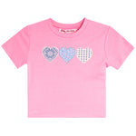A pink short-sleeve kids' t-shirt with three patterned heart graphics across the chest. The hearts feature blue and white patterns, including paisley, floral, and gingham.