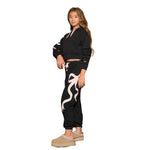 A young woman modeling a black two-piece tracksuit featuring a cropped hoodie with white drawstrings and white graphic details on the sleeves, paired with black jogger pants that have a large white ribbon graphic on the leg. She is wearing beige slip-on shoes against a solid white background.