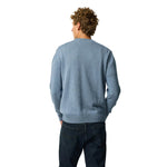 A man with curly brown hair is shown from behind wearing a light blue knit sweater and dark blue jeans against a solid white background.
