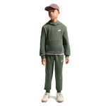A child model wearing a matching dark green Nike hoodie and sweatpants set, paired with a dusty rose baseball cap and white sneakers, standing against a plain white background.