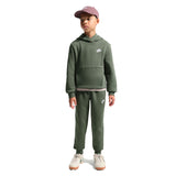 A child model wearing a matching dark green Nike hoodie and sweatpants set, paired with a dusty rose baseball cap and white sneakers, standing against a plain white background.