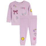 Two-piece set in light lavender purple, featuring a long-sleeve sweatshirt and matching pants. The set is decorated with cute cartoon graphics including a pink bow, a pink heart with wings, a yellow smiley face, a diamond, and a star w rainbow.