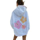 A woman shown from the back wearing a light heather gray hoodie with large colorful hibiscus flower graphics on the back in pink, yellow, and orange, plus a small multicolor pattern along the hood.