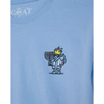 A close-up of a light blue t-shirt featuring a cartoon blue goat character wearing a gold crown and holding a menorah, with a white 'GOAT' brand logo printed inside the collar.
