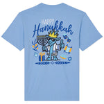 A light blue short-sleeve t-shirt shown from the back, featuring a colorful graphic of a cartoon dog wearing a crown and mezotall, surrounded by Hanukkah symbols including a menorah, dreidels, and gift boxes, with the text 'Happy Hanukkah'.
