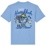 A light blue short-sleeve t-shirt shown from the back, featuring a colorful graphic of a cartoon dog wearing a crown and mezotall, surrounded by Hanukkah symbols including a menorah, dreidels, and gift boxes, with the text 'Happy Hanukkah'.