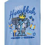 A light blue t-shirt featuring a colorful graphic design celebrating Hanukkah. The design includes the text 'Happy Hanukkah' in blue and white, a cartoon goat character wearing sunglasses and a crown holding a menorah, along with illustrations of a dreidel, matzah, gelto, and Star of David.