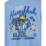 A light blue t-shirt featuring a colorful graphic design celebrating Hanukkah. The design includes the text 'Happy Hanukkah' in blue and white, a cartoon goat character wearing sunglasses and a crown holding a menorah, along with illustrations of a dreidel, matzah, gelto, and Star of David.