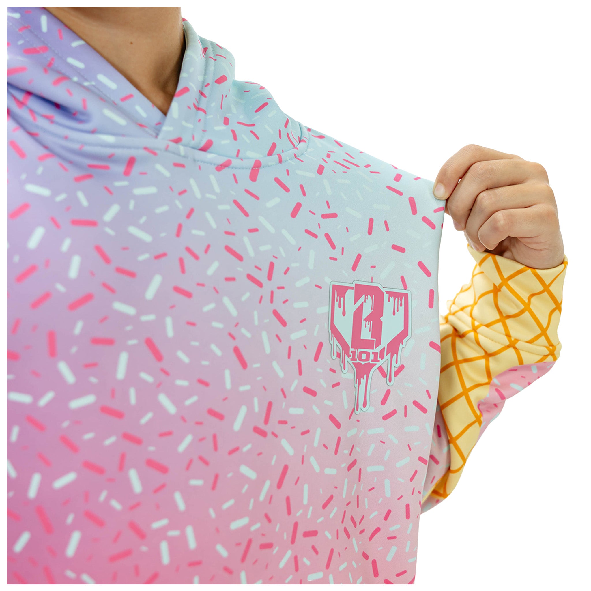 A close-up of a person wearing a hooded sweatshirt with a pastel gradient and sprinkle pattern. The hoodie transitions from light blue at the top to pink at the bottom, covered in pink and white sprinkle shapes.
