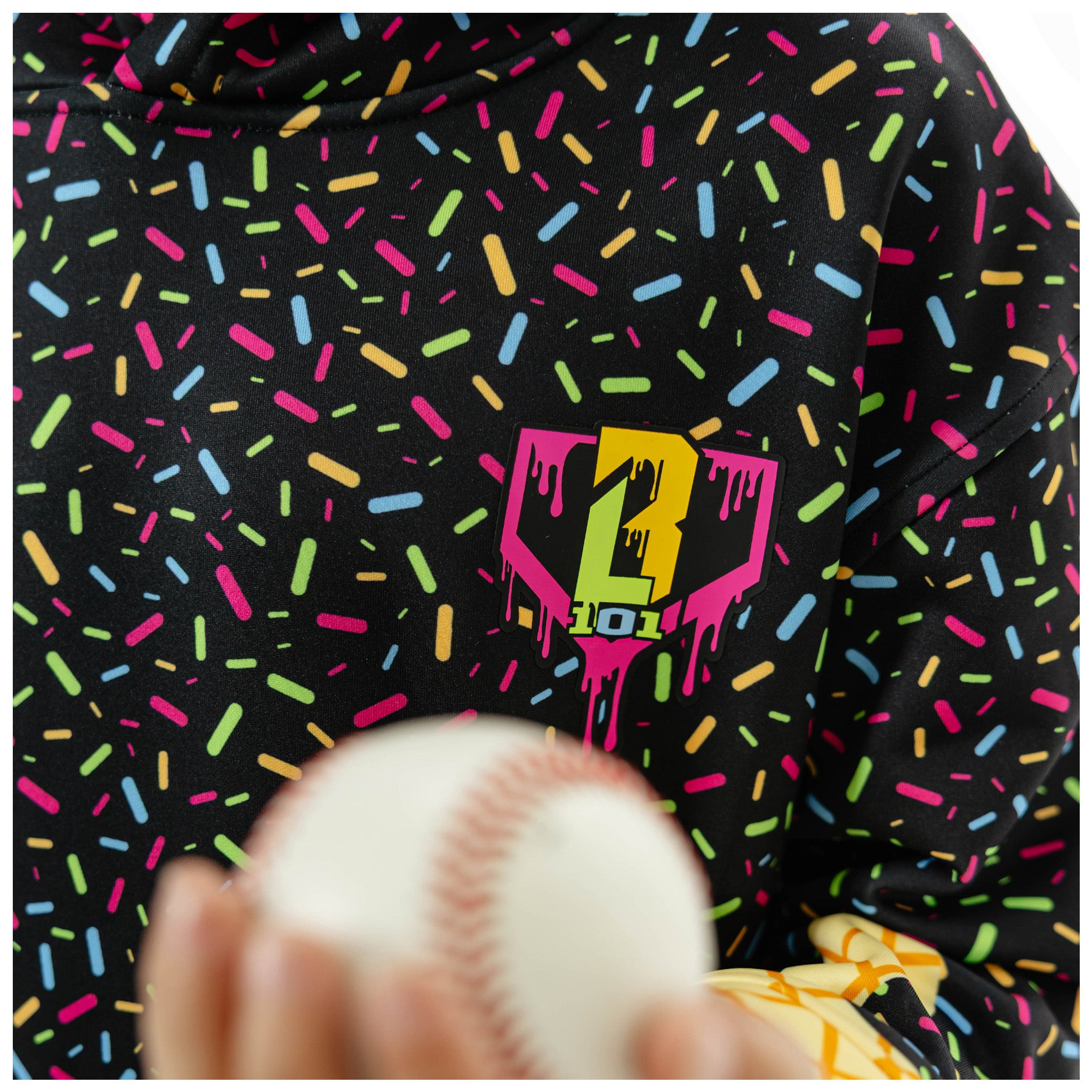 A close-up of a black hoodie covered in a colorful sprinkle pattern, featuring a neon pink and yellow dripping heart logo on the chest. A hand holding a baseball is visible in the foreground.