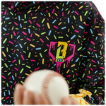 A close-up of a black hoodie covered in a colorful sprinkle pattern, featuring a neon pink and yellow dripping heart logo on the chest. A hand holding a baseball is visible in the foreground.