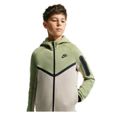 A boy wearing a Nike hooded zip-up jacket in sage green, beige, and black, photographed against a plain white background.