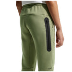 A close-up of a person wearing sage green Nike fleece jogger pants, showing the back pocket with a vertical black zippered pocket and a small black Nike swoosh near the lower leg.