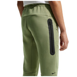 A close-up of a person wearing sage green Nike fleece jogger pants, showing the back pocket with a vertical black zippered pocket and a small black Nike swoosh near the lower leg.