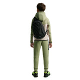 A child model shown from the back wearing a sage green and beige color-blocked hoodie, matching sage green sweatpants, neon green and black sneakers, and a black Nike backpack against a plain white background.