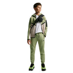 A child model wearing a matching Nike tracksuit in sage green, featuring a zip-up hoodie with beige and green color-blocked panels, matching sweatpants, a white t-shirt, and a black crossbody bag, set against a plain white background.