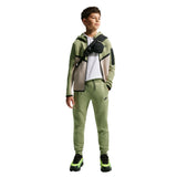 A child model wearing a matching Nike tracksuit in sage green, featuring a zip-up hoodie with beige and green color-blocked panels, matching sweatpants, a white t-shirt, and a black crossbody bag, set against a plain white background.