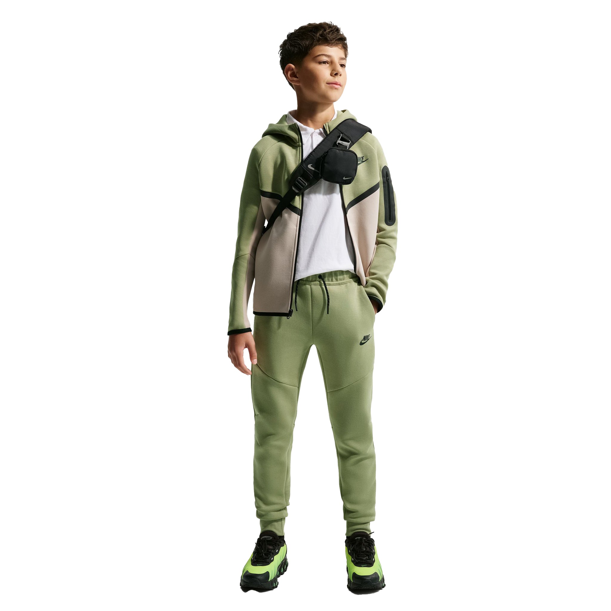 A child model wearing a matching Nike tracksuit in sage green, featuring a zip-up hoodie with beige and green color-blocked panels, matching sweatpants, a white t-shirt, and a black crossbody bag, set against a plain white background.