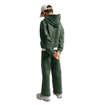 A child shown from the back wearing a dark green cropped hoodie and matching sweatpants, a white baseball cap, and white sneakers, against a plain white background.