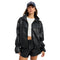 A model wearing a black faux-leather zip-up jacket with flap chest pockets, styled with black shorts, a white crop top, and a navy blue baseball cap with white text, set against a plain white background.