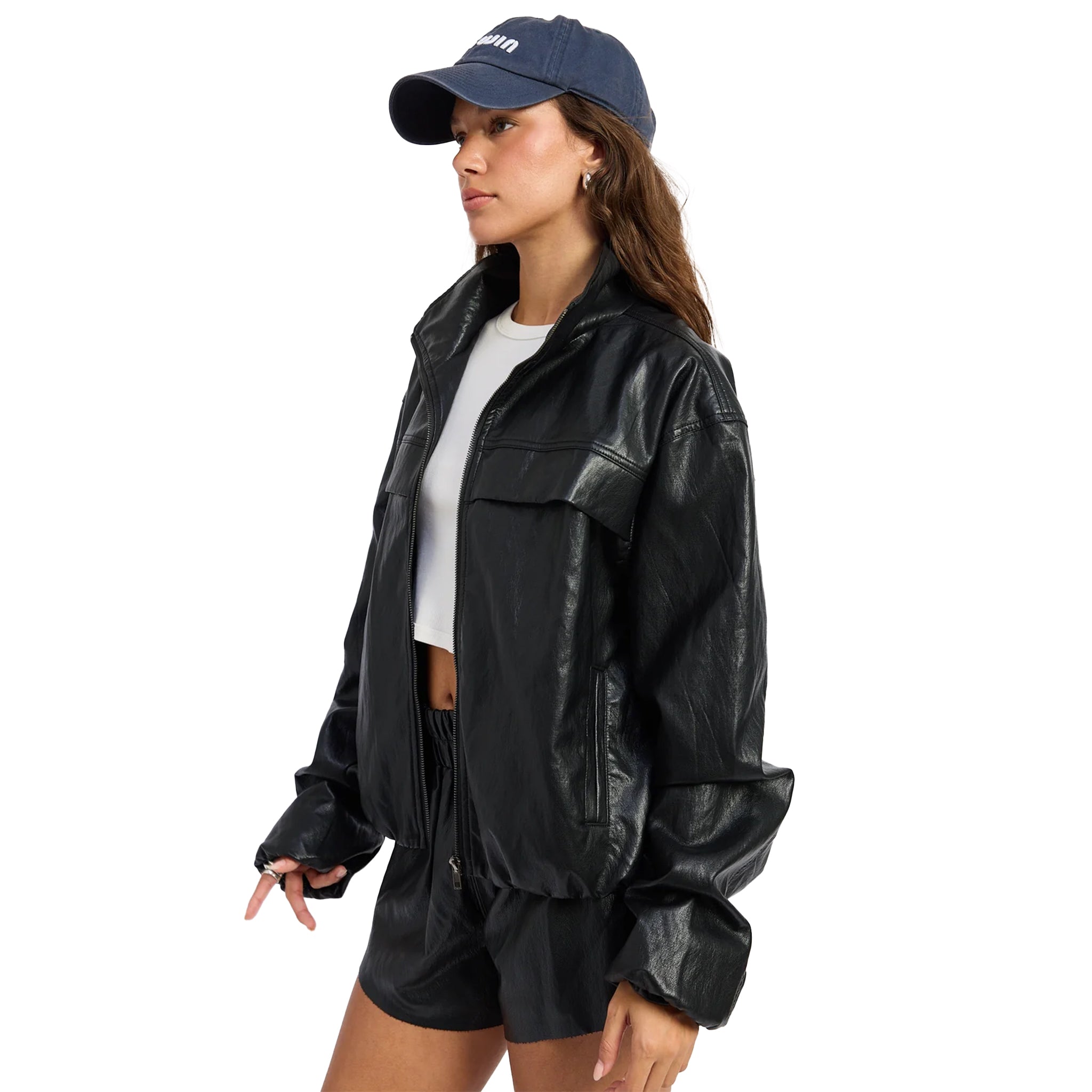 A woman wearing a black faux-leather jacket, black shorts, a white crop top, and a navy blue baseball cap, shown from a side profile against a solid white background.