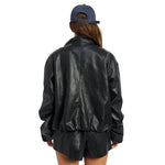 A person shown from the back wearing a black faux-leather jacket, black shorts, and a blue baseball cap against a plain white background.