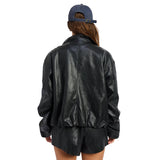 A person shown from the back wearing a black faux-leather jacket, black shorts, and a blue baseball cap against a plain white background.
