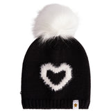 A black knit beanie hat featuring a fluffy white faux-fur pom-pom on top and a white heart-shaped knit motif on the front. A small white fabric tag with a gold star logo is visible on the lower right edge.