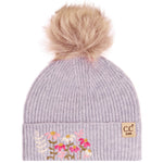 A light lavender ribbed knit beanie hat with a fluffy tan pom-pom on top and floral embroidery on the front cuff. A small tan brand patch reading 'c.