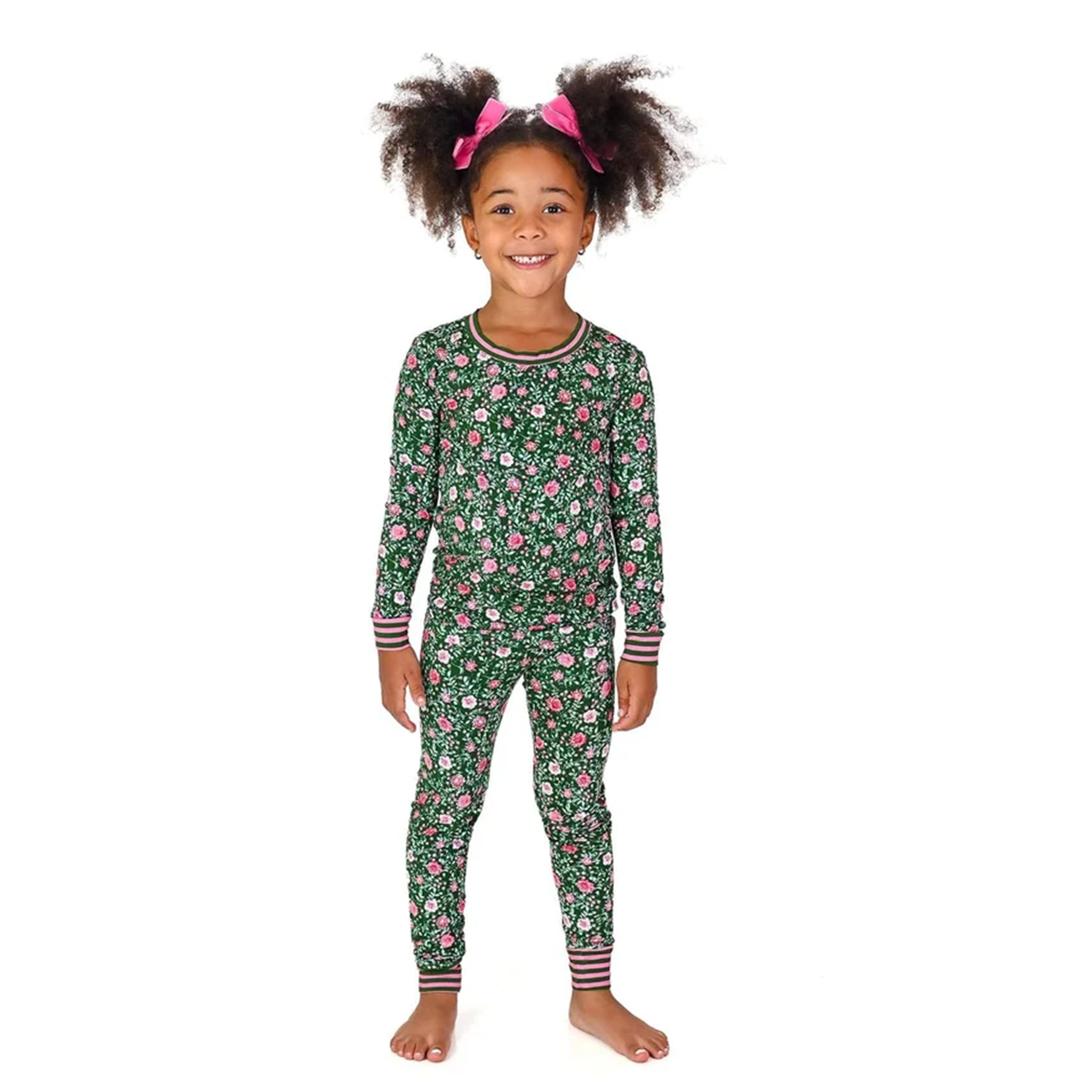 A young girl with dark curly hair in pigtails wearing a green long-sleeve pajama set with a pink and white floral pattern, standing against a solid white background.
