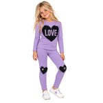 A young girl modeling a lavender purple two-piece set with black sequined heart patches on the chest and knees. The long-sleeve top features a large black heart with the word 'LOVE' in white sequins.