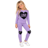 A young girl modeling a lavender purple two-piece set with black sequined heart patches on the chest and knees. The long-sleeve top features a large black heart with the word 'LOVE' in white sequins.