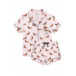 A two-piece women's pajama set featuring a short-sleeve button-front top and matching shorts in light pink, printed with an all-over dachshund pattern and finished with black piping and a black drawstring.
