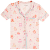 A short-sleeve pajama shirt in pale pink with a watercolor-style print of cakes, and bows, featuring light blue piping and a button-front collar.