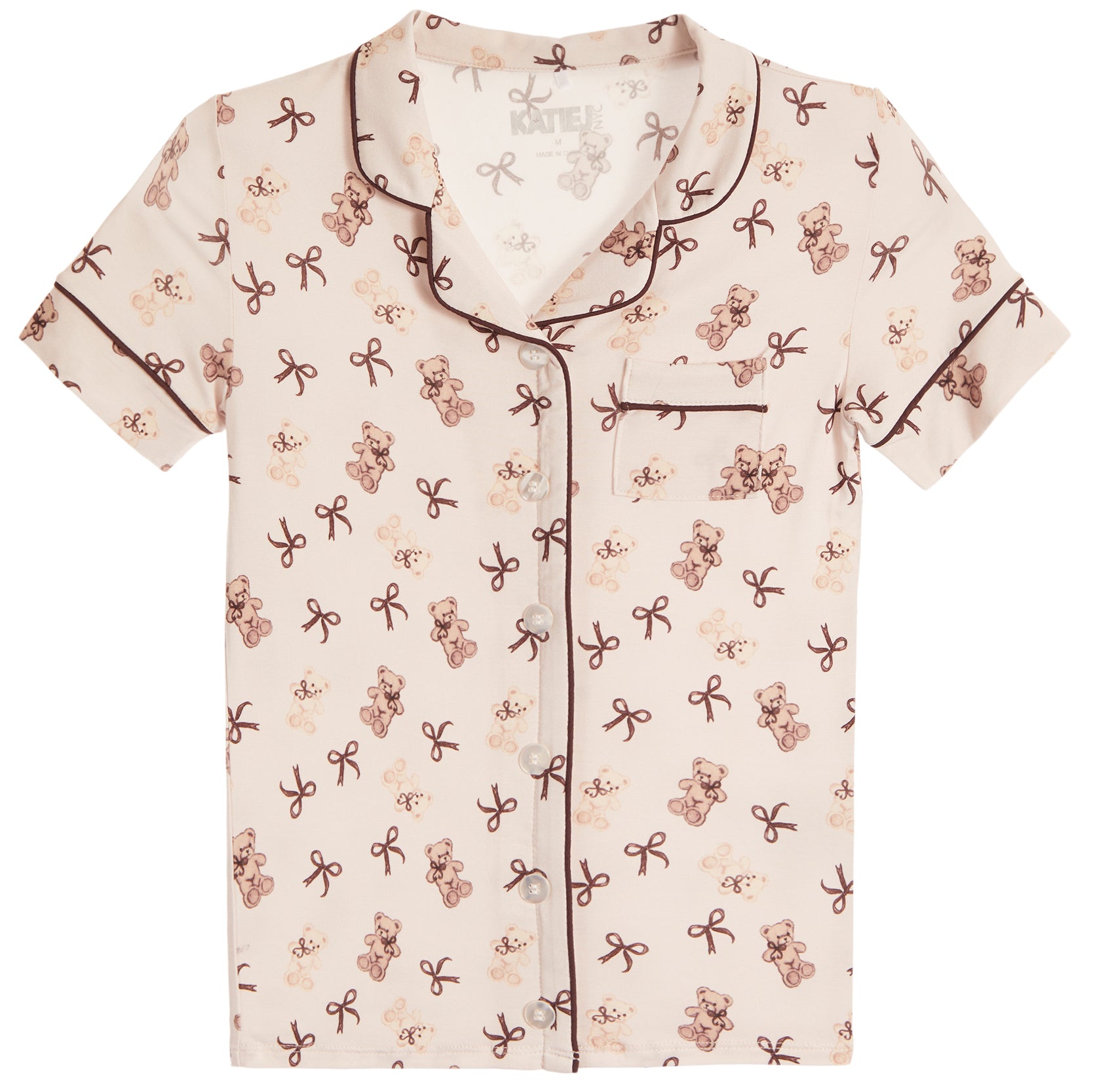A short-sleeve pajama shirt with an all-over teddy bear and bow print on a warm cream background, featuring dark brown piping along the collar, placket, cuffs, and chest pocket.