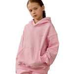 A young girl wearing a light pink oversized hoodie sweatshirt with a small embroidered  logo on the chest, set against a plain white background.