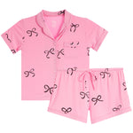 A two-piece baby pajama set in pink, featuring a short-sleeve button-front top with a chest pocket and matching shorts, both decorated with a black bow pattern, shown on a plain white background.