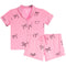 A two-piece baby pajama set in pink, featuring a short-sleeve button-front top with a chest pocket and matching shorts, both decorated with a black bow pattern, shown on a plain white background.