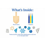 An informational product shot titled "What's Inside:" showing the contents of a craft kit on a white background. The kit includes a wooden dreidel, three markers in blue, yellow, and light blue, several blue and silver glitter mosaic tiles, sparkly gems, and small silver no-mess glue dots.