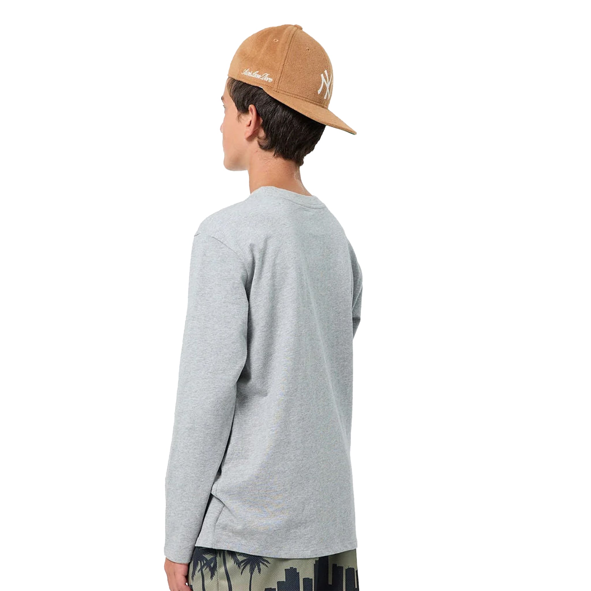 A boy from the back wearing a light gray long-sleeve t-shirt, a tan baseball cap with a white embroidered logo, and dark patterned shorts, set against a plain white background.