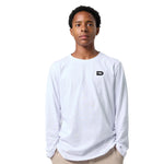 A young man wearing a white long-sleeve crewneck t-shirt with a small black stylized 'D' logo on the left chest, paired with beige pants, shot against a solid white background.