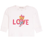 A white long-sleeve baby/toddler top featuring a central graphic of a teddy bear wearing a pink bow and jeans, sitting on the word 'LOVE' in pink letters.