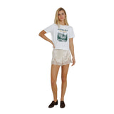 A woman modeling a white graphic t-shirt with a Jackson Hole mountain print, paired with cream lace-trimmed shorts and dark brown loafers, against a solid white background.